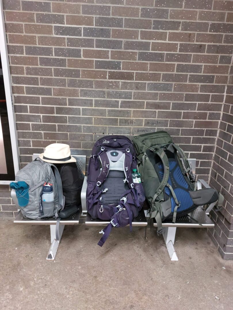 Two daypacks and two big Osprey backpacks on a metal bench