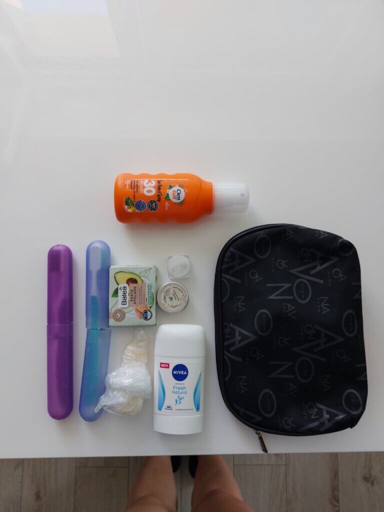 Cosmetics for a trip laid out on a table. There is a bottle of sunscreen, two toothbrushes, solid conditioner bar, solid deodorant, solid perfume, mini cream, soap wrapped in a plastic bag and a cosmetics bag.