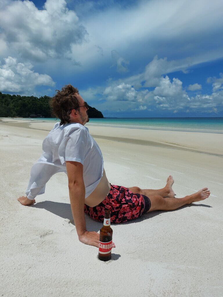Daniel sitting on a beach with a Cambodia beer stuck in the sand next to him
