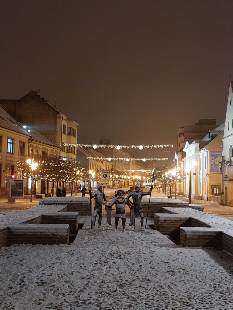 Snow-dusted pedestrian zone with Christmas lights and statues of knights and a herald