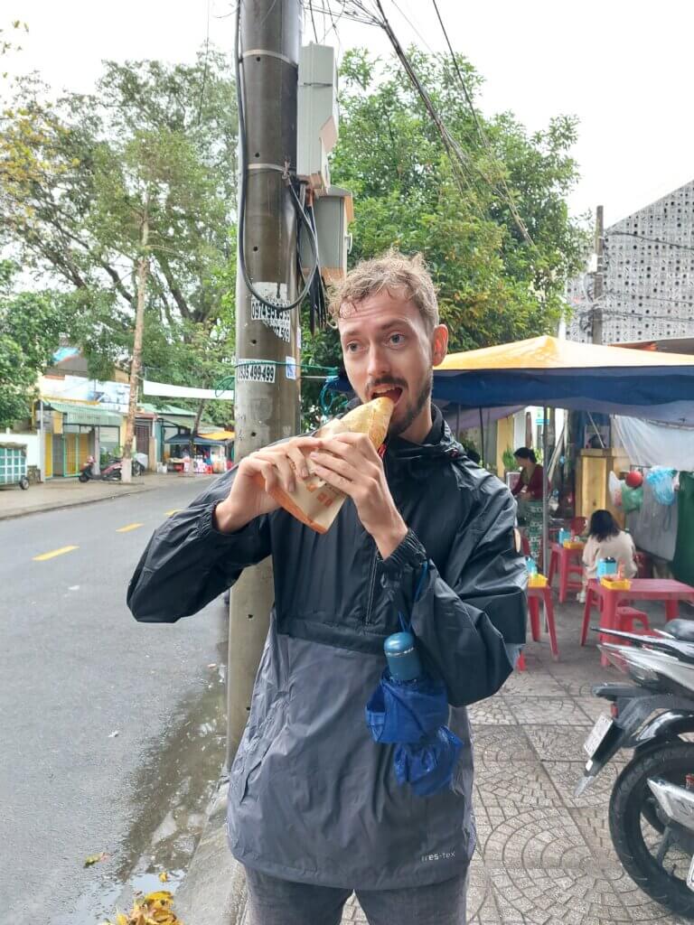 Daniel standing on a street in Vietnam wearing a rain jacket and eating a banh mi sandwich