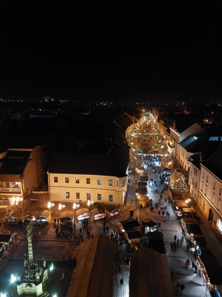 Nighttime shot of a part of a main square and a pedestrian zone, all decorated for Christmas and with wooden huts of the Christmas market spread around