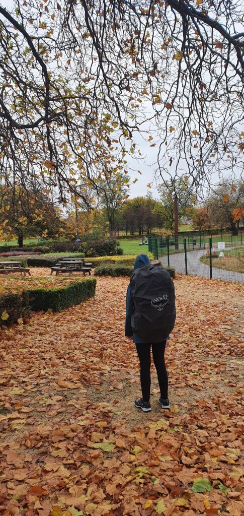 Mirka standing in a park during autumn, with leaves on the ground everywhere. It's raining so she is wearing a rain jacket and her big Osprey backpack with a raincover on. She is facing away from the camer