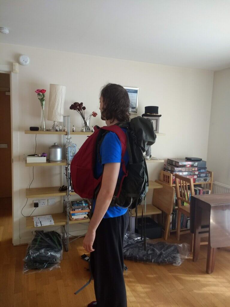 Daniel standing in a living room with a day backpack on his front and a big backpack on his back with more backpacks lying on the floor