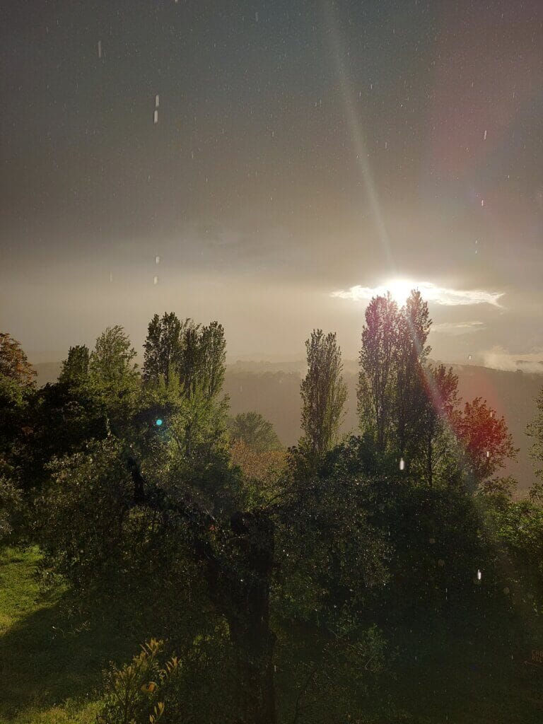 A view of Italian countryside with sun shining through clouds but also rain droplets falling in front of the camera lens.
