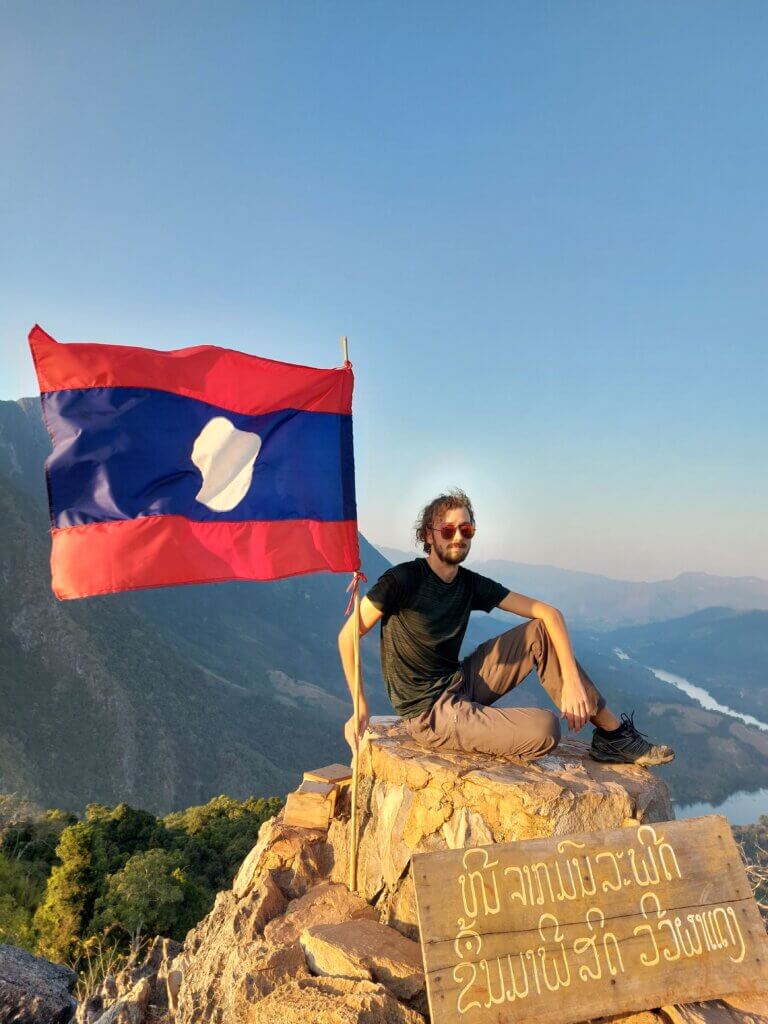 Daniel, sitting on top of a rocky peak with views of Laos behind him, holding the Lao flag.
