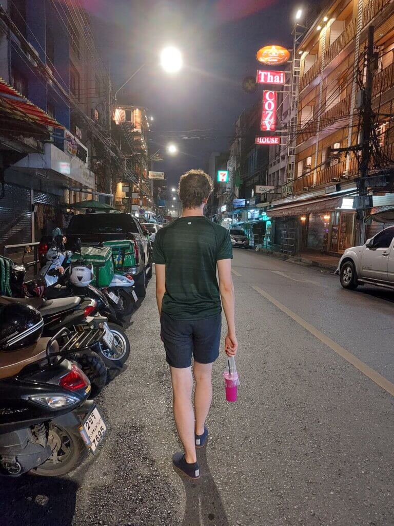 Daniel walking along a road of night time Bangkok carrying a pink dragon fruit smoothie in his right hand