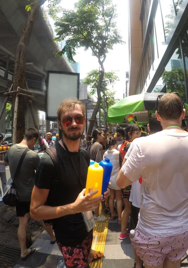 Daniel standing on a busy street with other people celebrating Songkran, holding two empty water gun tanks.