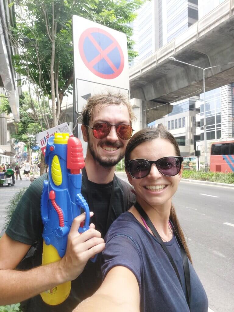 Daniel and Mirka standing on a street during Songkran with Daniel holding up a water gun. Both are wearing sunglasses.