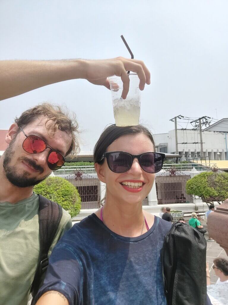 Mirka and Daniel taking a selfie in Wat Arun with Daniel holding a cup of ice-cold coconut water on Mirka's head to cool her down