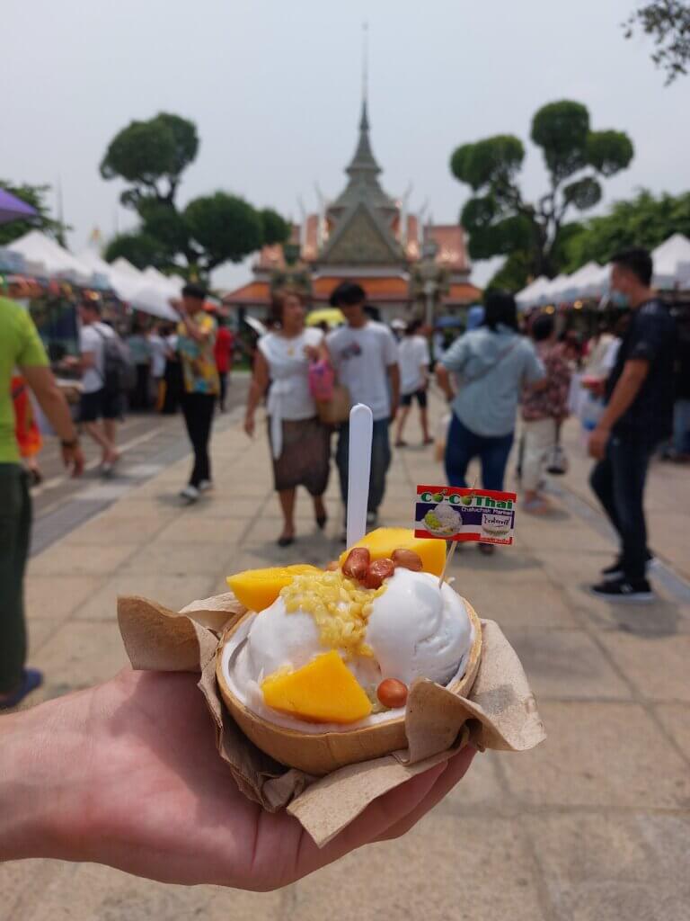 A bowl of coconut ice cream with mango, peanuts and rice krispies held against a background of people in Wat Arun during Songkran