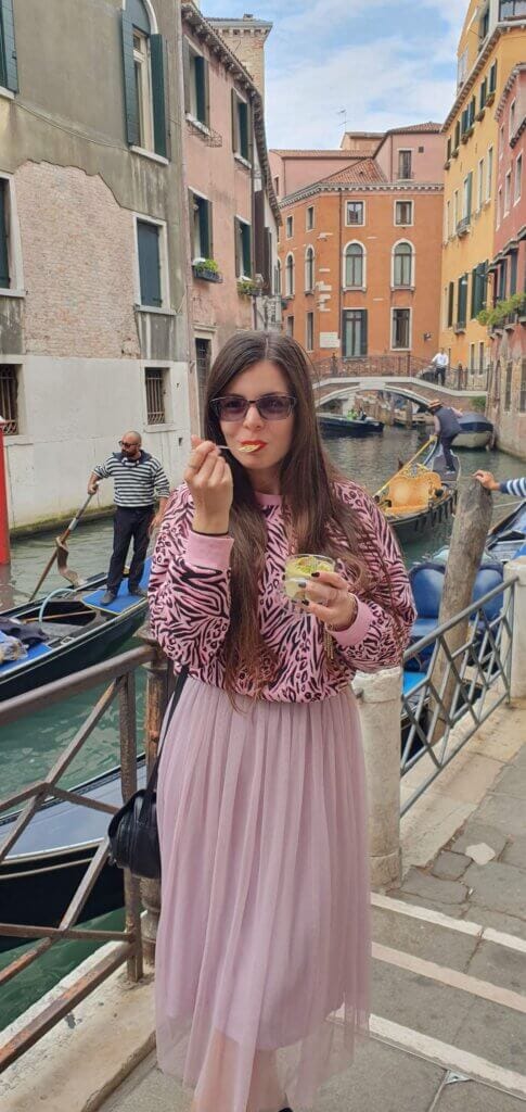 Mirka standing next to a canal in Venice with gondolas behind her, eating a tub of tiramisu