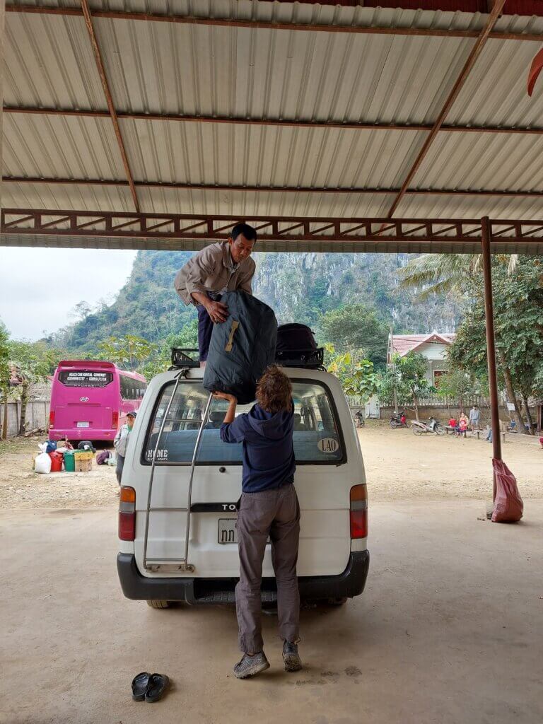Daniel passing a big backpack to a man standing on the roof of a van to store it there during our journey