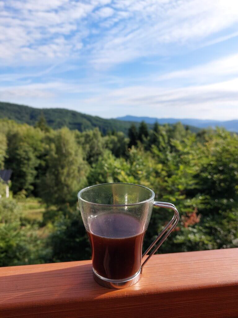 A glass of coffee on a wooden railing overlooking trees and mountains