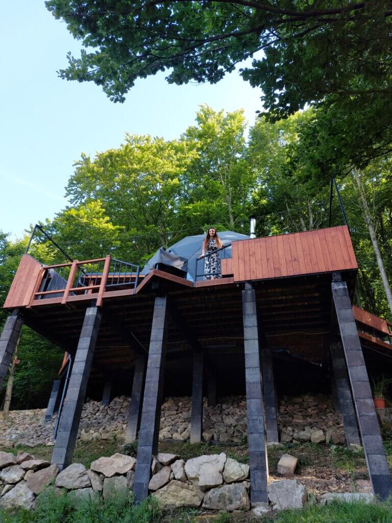 A wooden terrace on high pillars with the geodome at the top and Mirka leaning over the railing