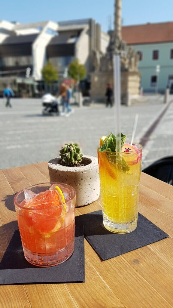 Two cocktails on a table with a blurry town square in the background