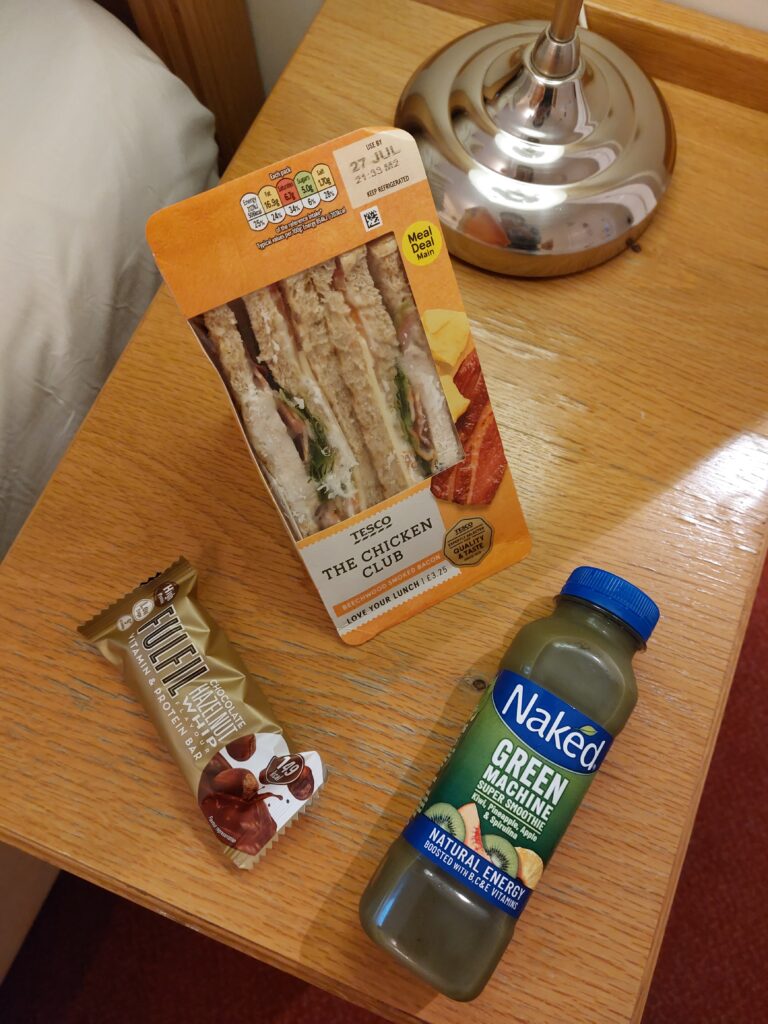 Sandwich, protein bar and a juice bottle on a bedside table in a hotel room