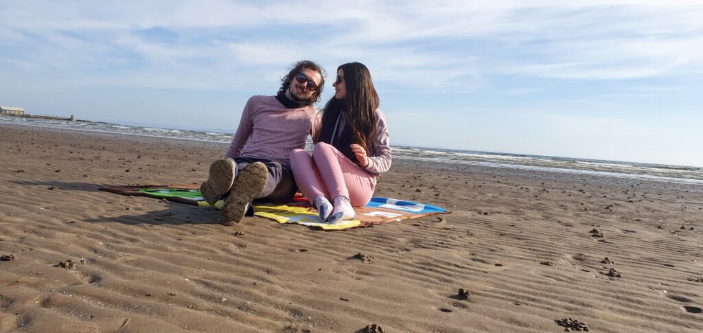Mirka and Daniel sitting on a blanket on an empty beach with the sea behind them