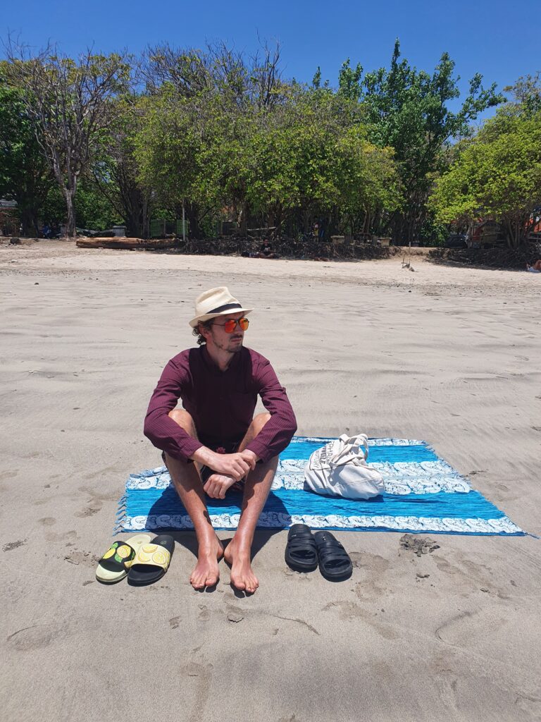 Daniel sitting on a beach on a big blue sarong