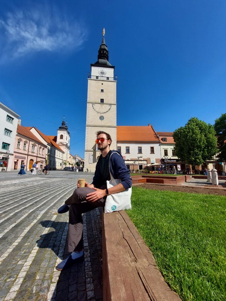Daniel sitting beneath a town tower holding a cup of coffee